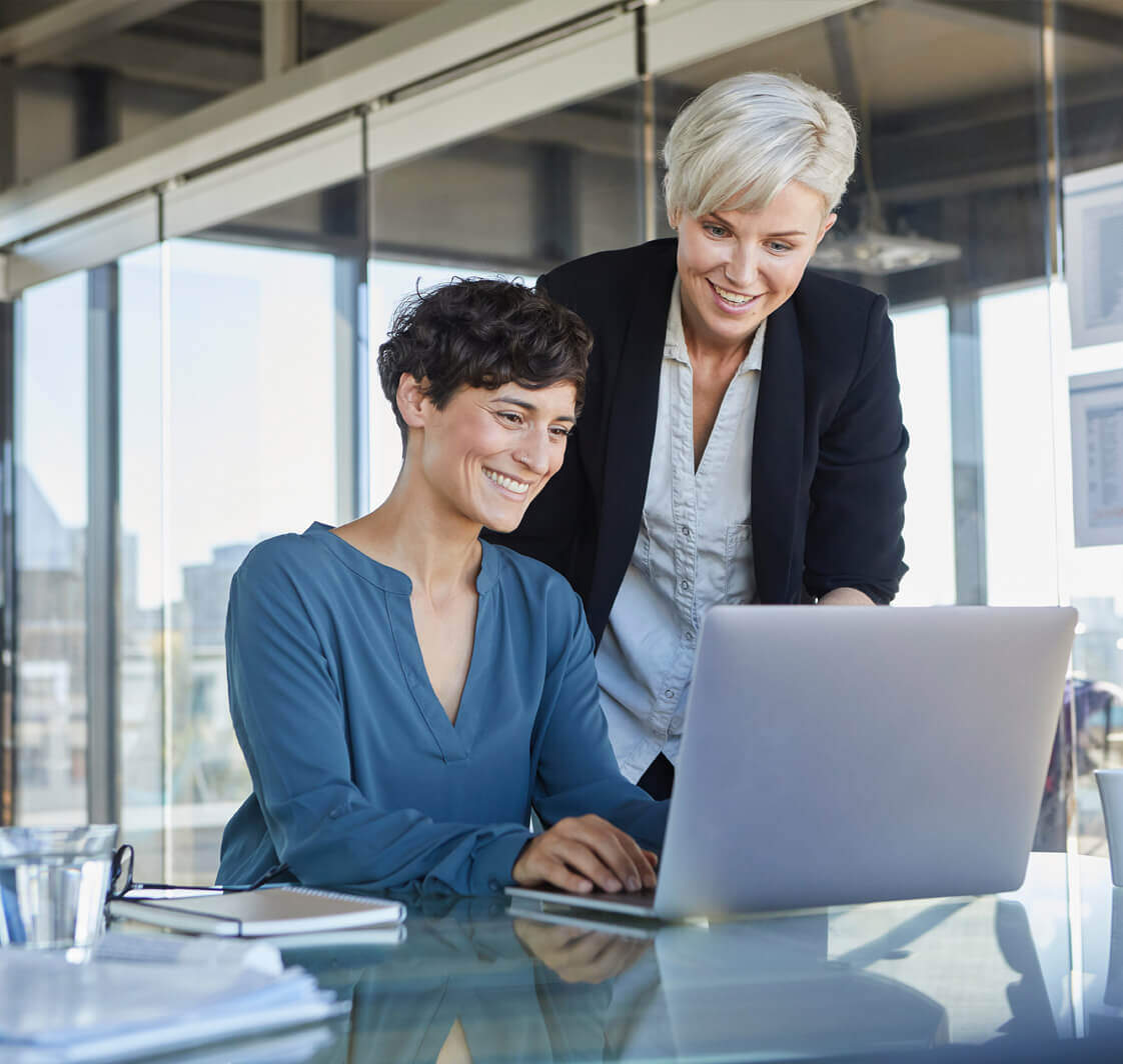 Two smiling businesswomen sharing laptop at desk in office.