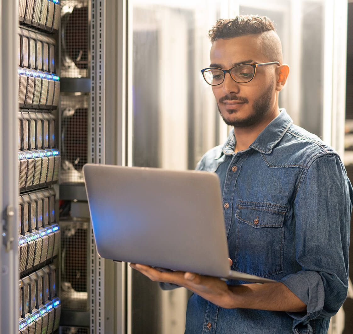 Serious skilled young Arabian IT specialist with beard standing by modern server and using laptop while monitoring network processes