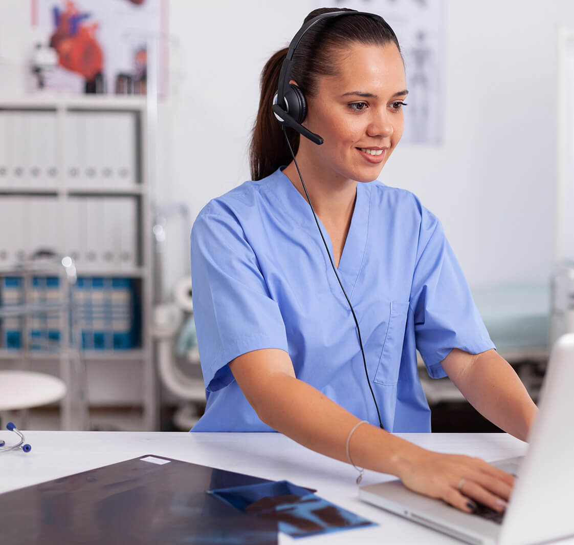 Medical receptionist wearing headset with microphone in private hospital typing on laptop.