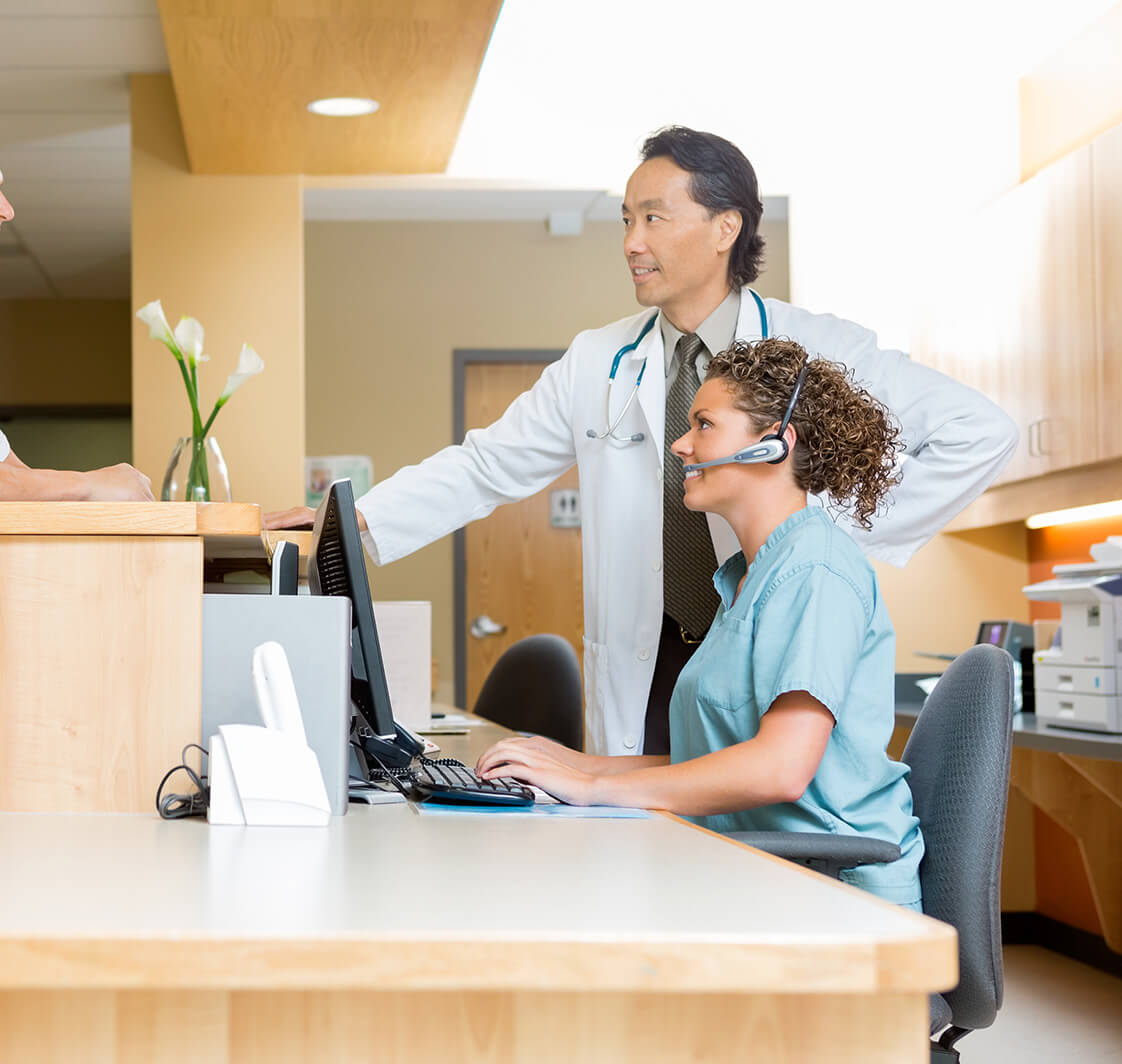 Patient With Doctor And Nurse At Reception Desk.