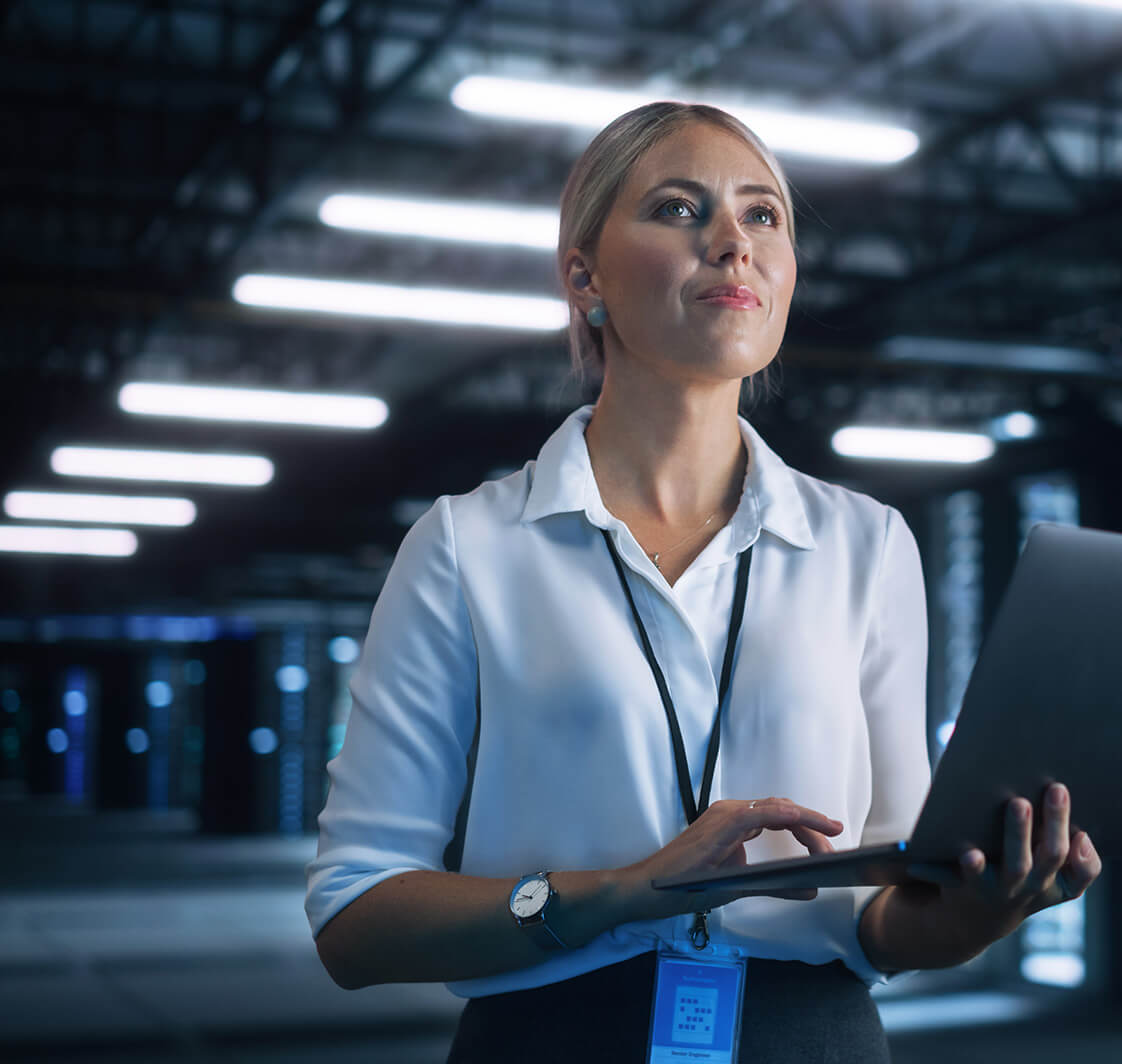 Female Data Center IT Technician Standing at the Server Rack Corridor with Laptop Computer. She is Visually Inspecting Something while Looking at the Screen. Night Office Concept.