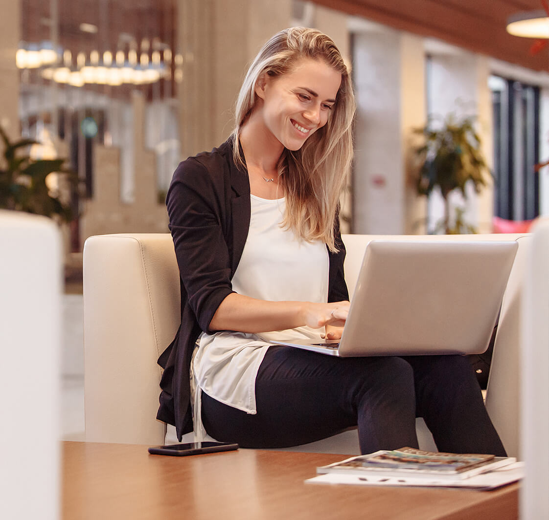 Laughing young beautiful blonde woman is sitting in lobby of hotel and waiting for check-in checking mail and social networks using a laptop and high-speed Internet.