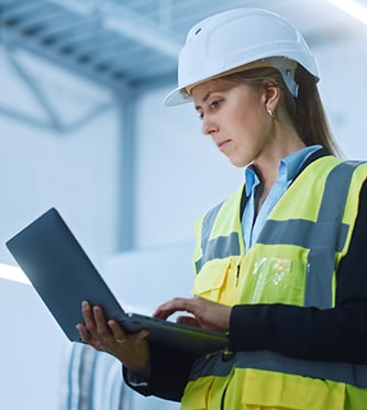 High-Tech Factory: Confident and Professional Female Engineer Wearing Safety Jacket and Hard Hat Holding and Working on Laptop Computer. Modern Bright Industrial Facility.