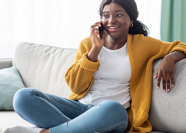 Young woman, sat on a sofa, smiling and talking on a mobile handset.
