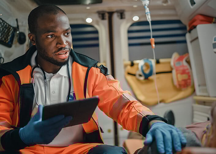 A male ambulance driver in the driver's seat in an ambulance, with a stethoscope around his neck, and wearing a hi-vis jacket.
