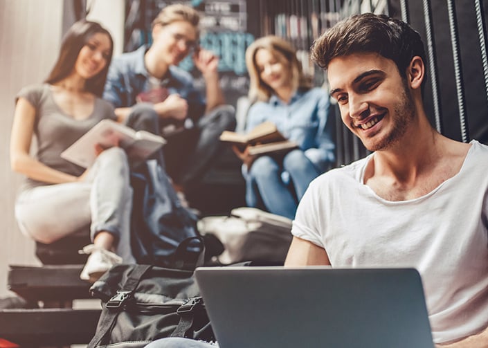 Group of students in higher education, sitting on the stairs together, smiling, and reading books and screens. 