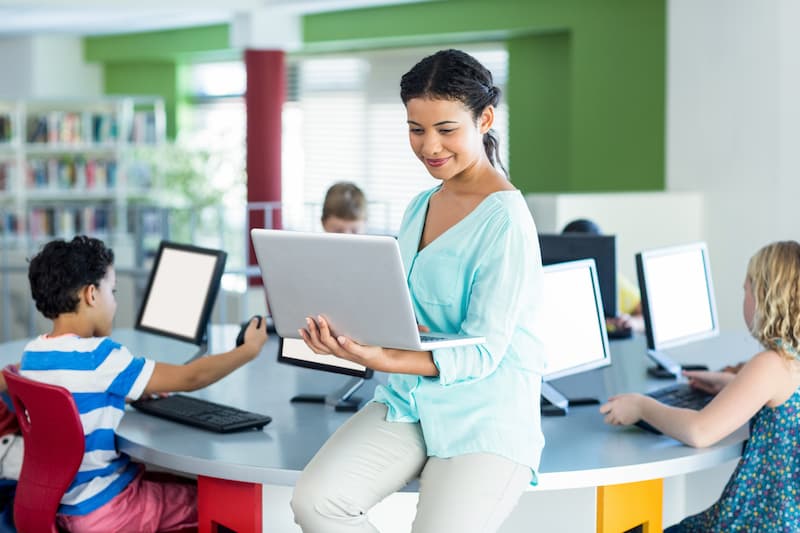 Teacher in a classroom leaning on a desk smiling, with her laptop