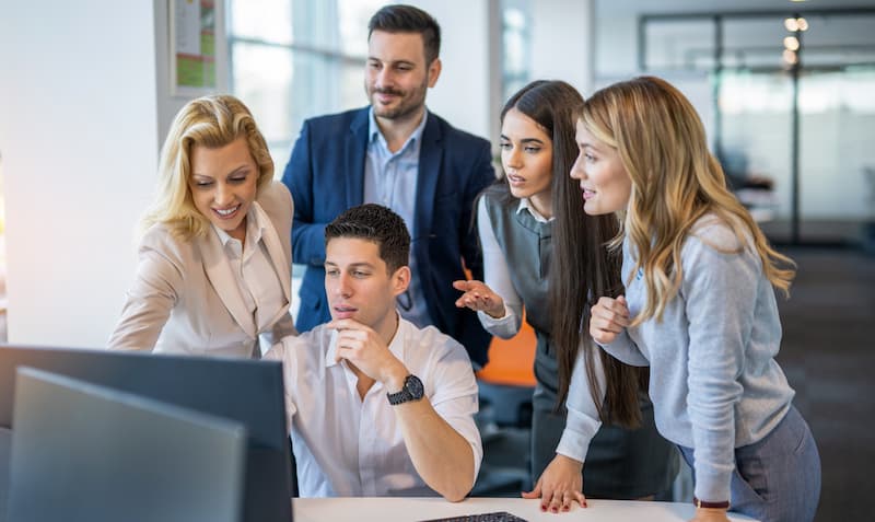 A group of office workers smiling stood around a laptop