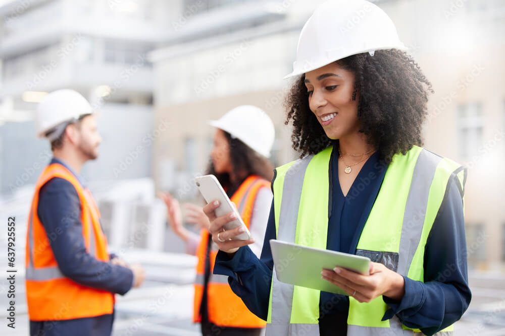 A construction worker in a hard hat and hi-vis jacket looking at her mobile phone and holding a tablet on site with other construction workers