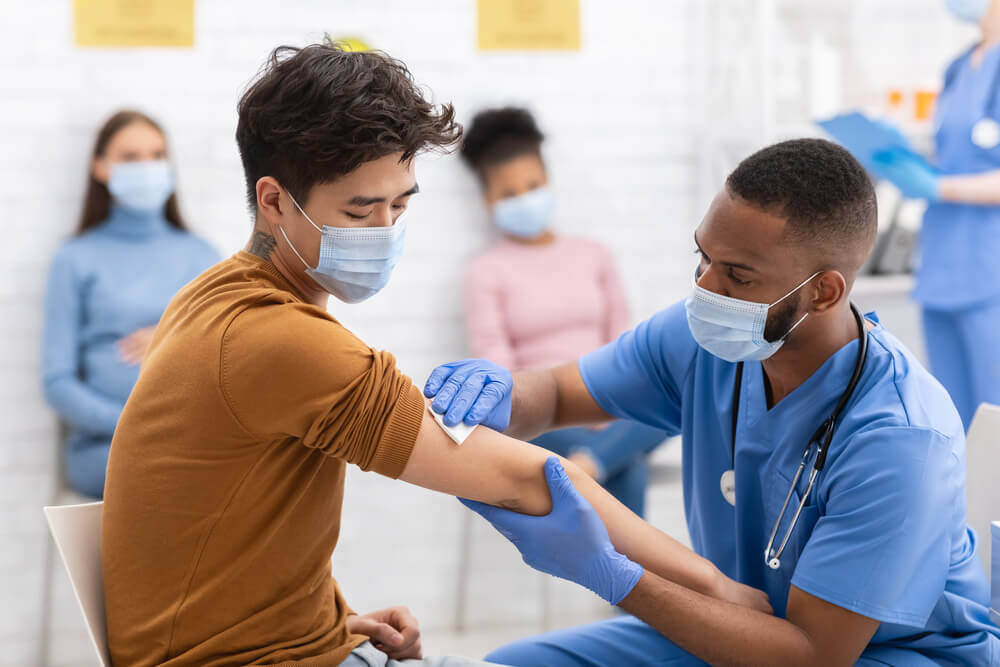 A male nurse in blue scrubs and a face mask is preparing a male patient's arm for a vaccine, and in the background are female patients sat waiting, as part of the national vaccine roll-out.